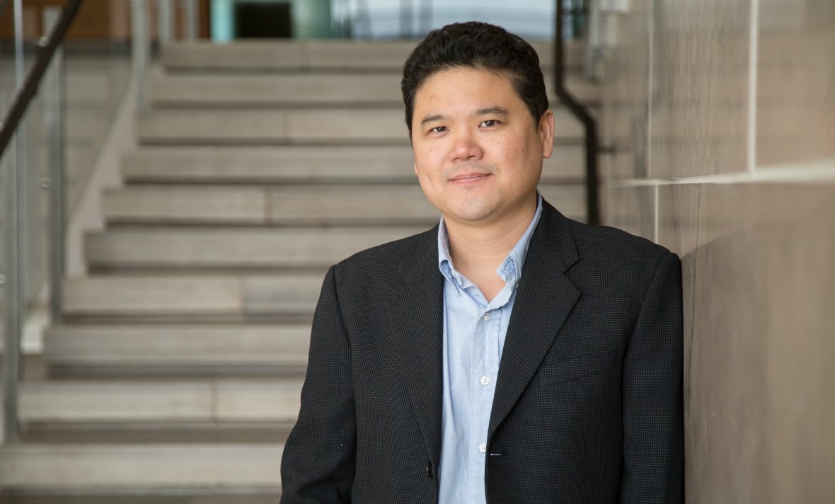 As Asian man wearing a black suit jacket and blue dress shirt leans against a wall in front of a set of stone stairs. 
