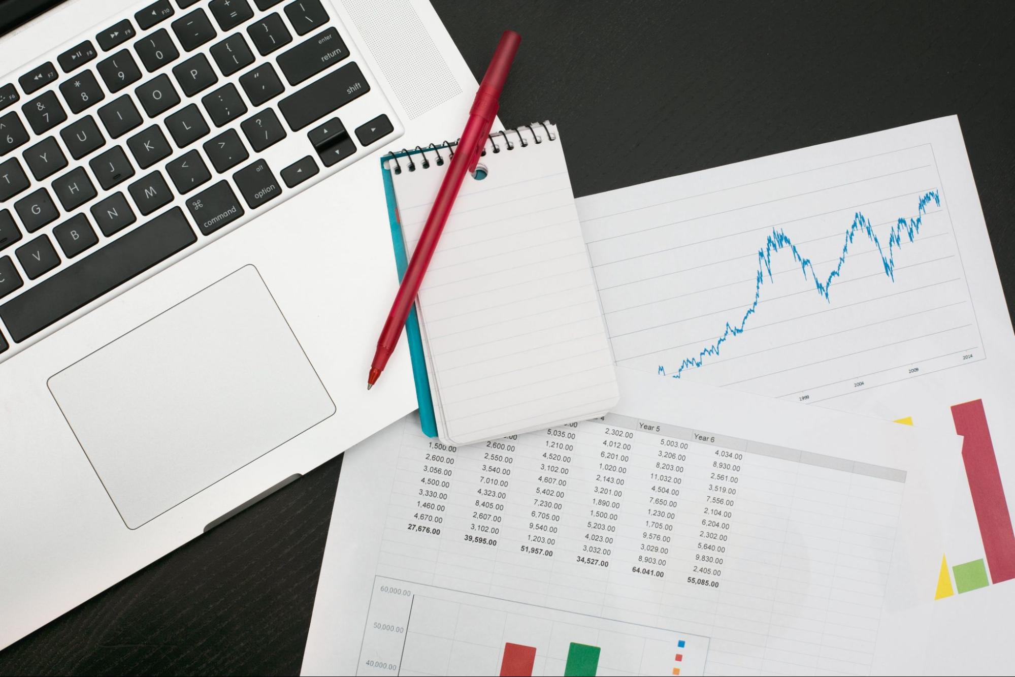 Flatlay of a red pen, notepad, laptop, and business papers with numbers and graphs. These are necessary tools to track finances.
