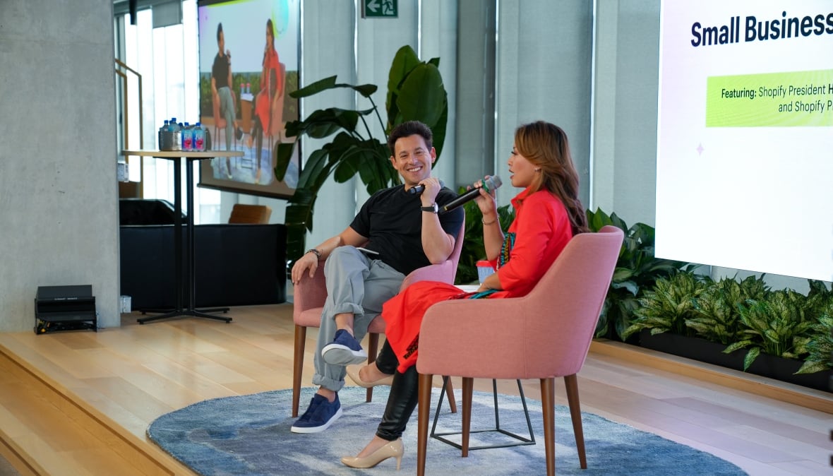 A woman in bright red coat sits in a modern chair holding a microphone speaking with a man in jeans and a t shirt. They both are on a stage with a projector screen that says small business behind them. 