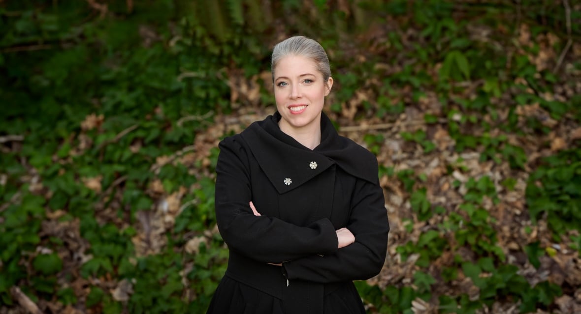 A woman with grey blond hair in a ponytail wearing a black wool jacket with fancy silver buttons and a large lapel stands with her arms crossed in front of wall covered in green and brown fall foliage. 