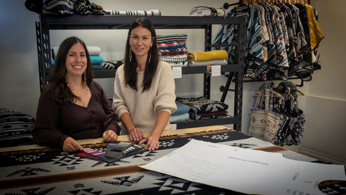 Two women stand near a large table covered by a blanket featuring Indigenous designs like an eagle. Ponchos and bags hang in the background.
